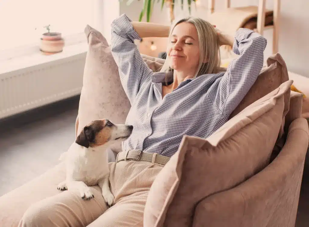 Content middle-aged woman relaxing on couch with her dog, feeling emotionally well with the support of DHEA supplementation.